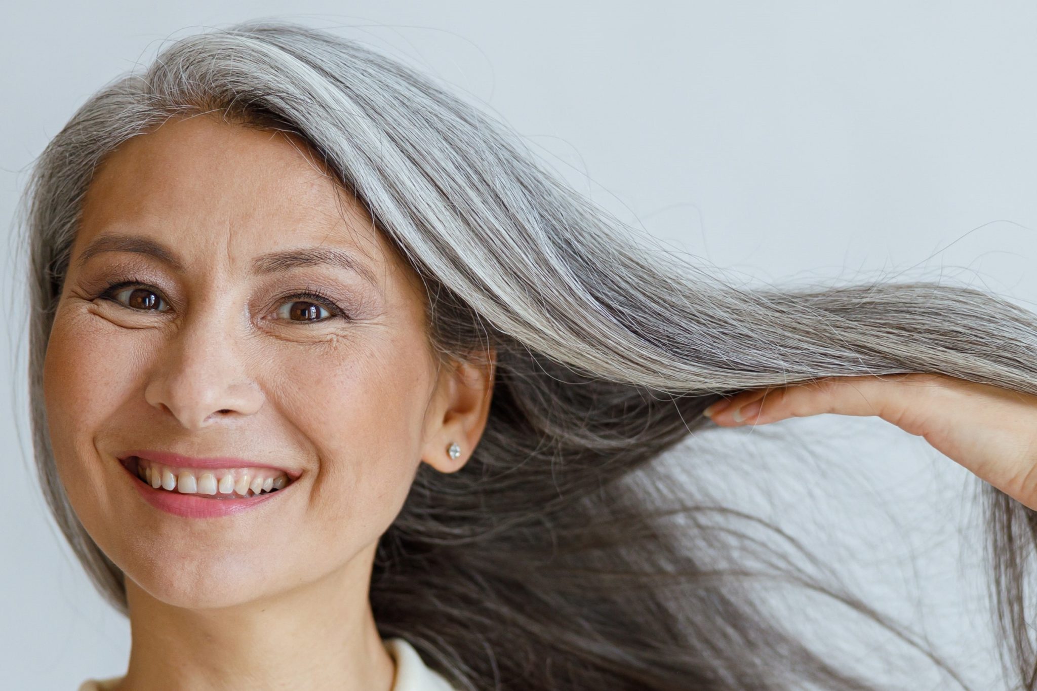 Happy middle aged Asian lady shows natural silver hair posing on light background in studio. Mature beauty lifestyle
