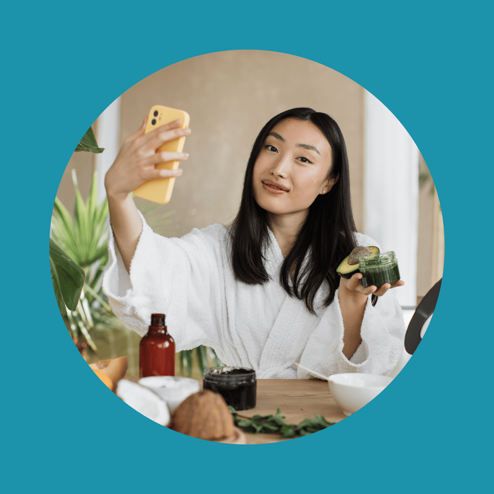 Asian brunette shoulder length hair female social media influencer in bathrobe streaming and introducing beauty products to audience with phone holding glass jar with cream, skin care concet.